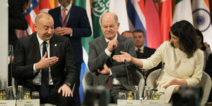 Ilham Alijew, Olaf Scholz und Annalena Baerbock sitzen nebeneinander auf einer Konferenz, im Hintergrund die Flaggen verschiedener Länder